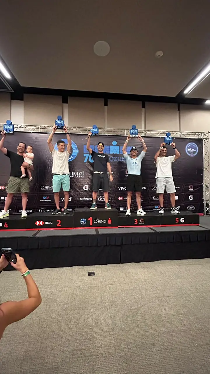 Pódio da categoria no IRONMAN 70.3 Cozumel 2026, em frente ao painel de patrocinadores